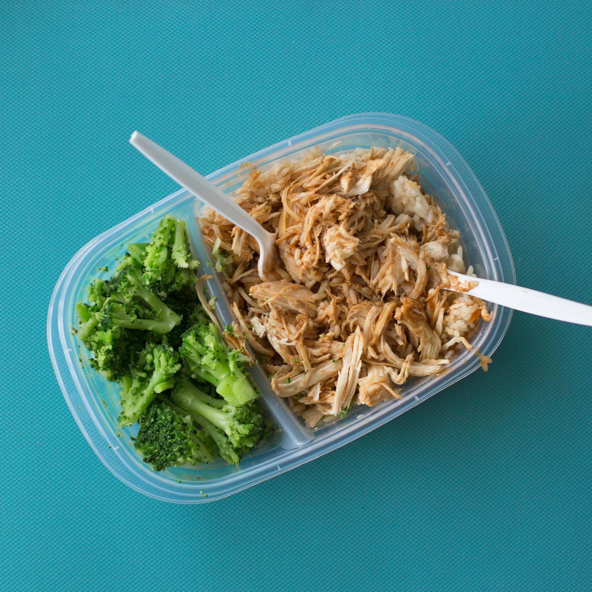 Meal prep container with broccoli and shredded chicken, set on a blue surface, with a white plastic fork.
