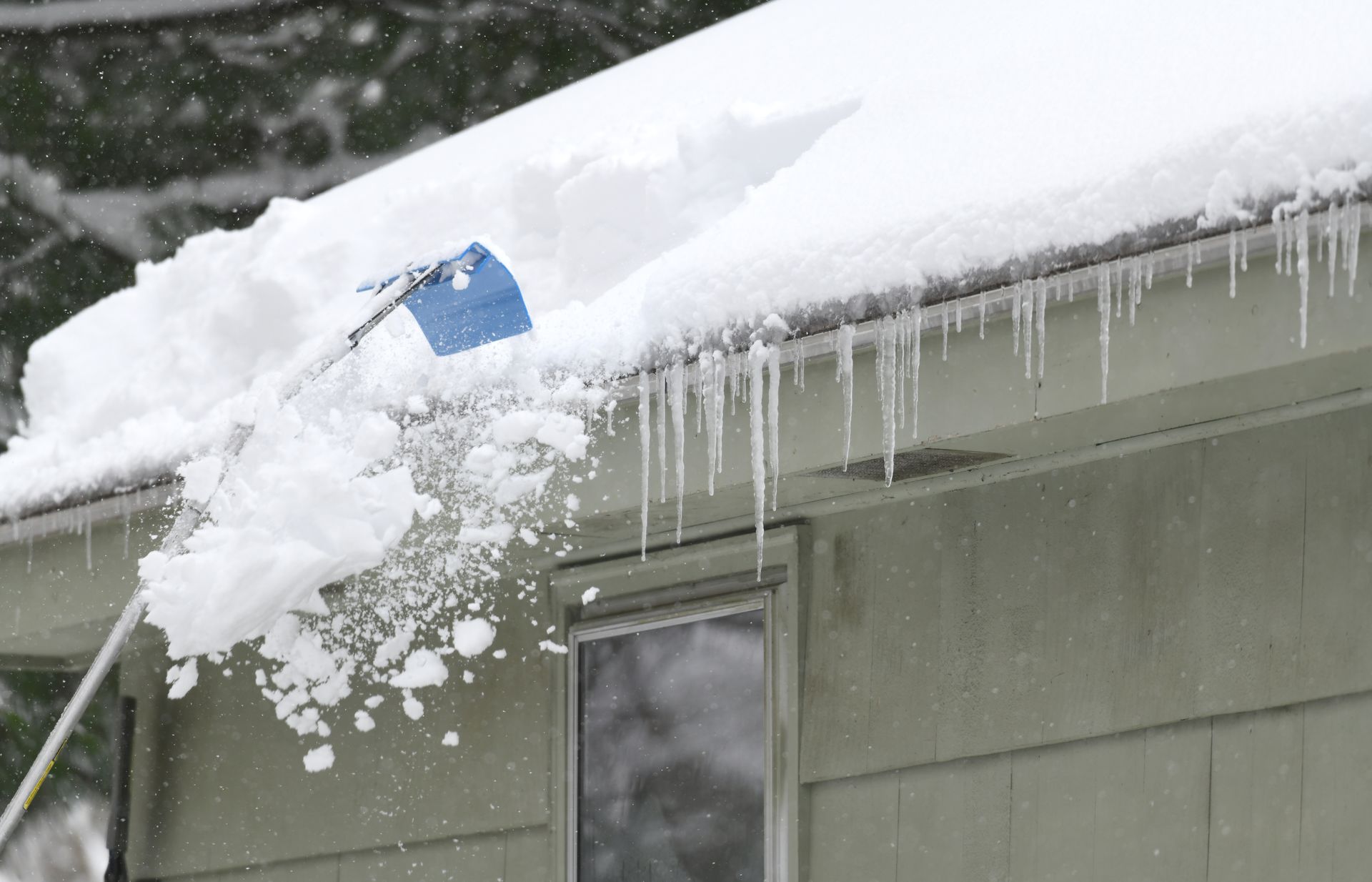 Shoveling snow off a roof with a long-handled tool. Icicles hang from the eaves.