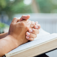 A person is praying with their hands folded over a bible.