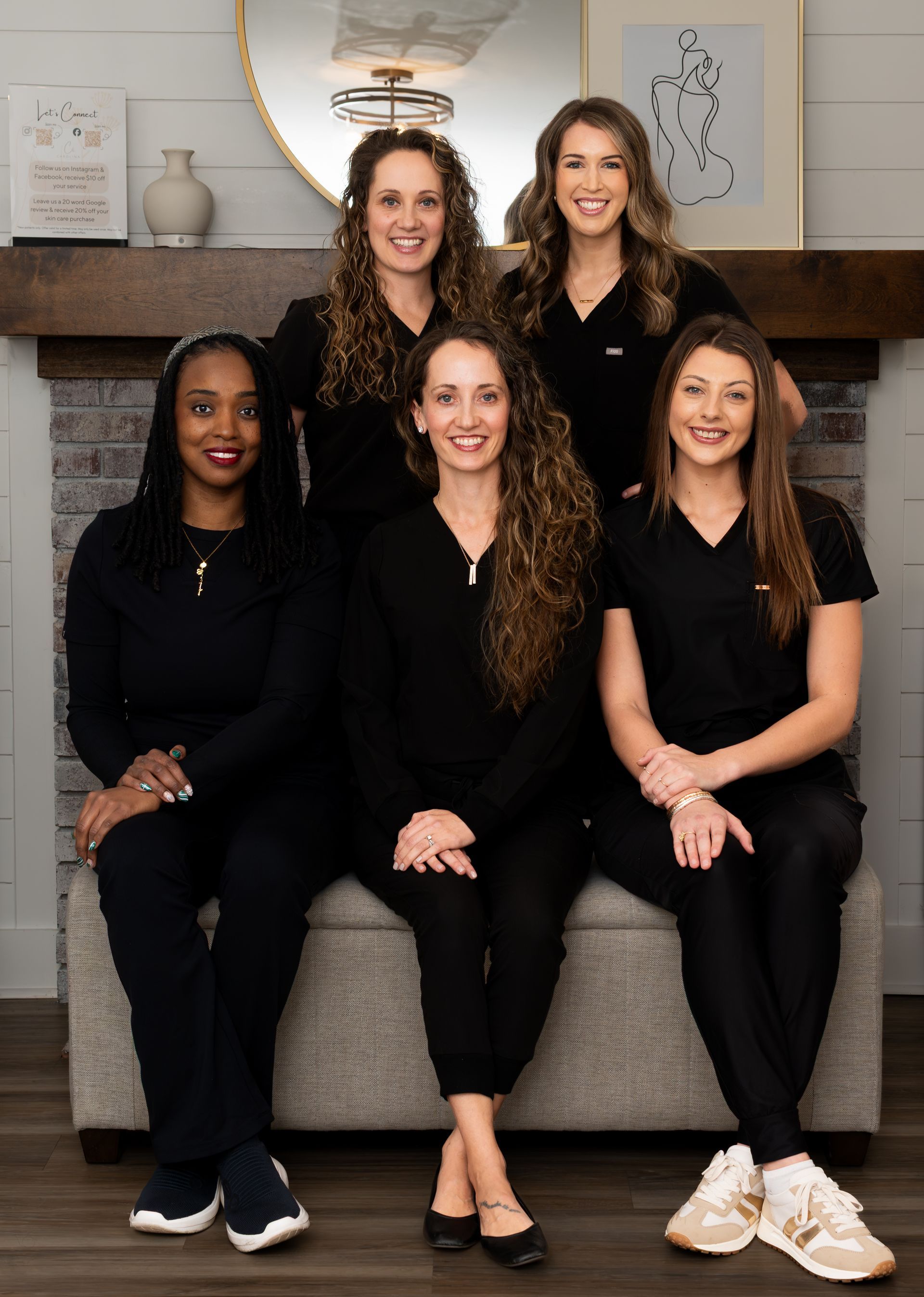 Five women in black outfits posed together on a couch in a living room by a fireplace.