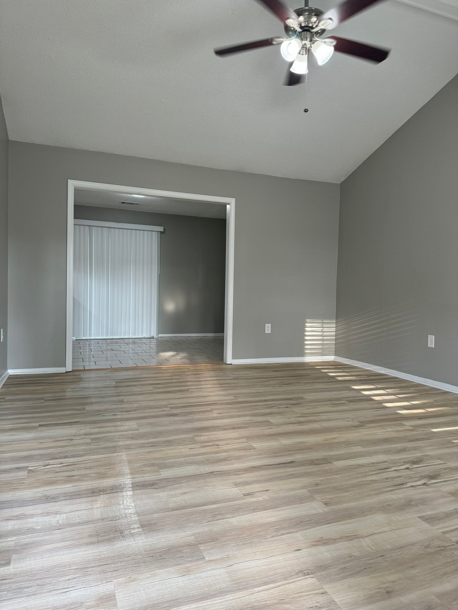Empty room with gray walls and wood-look flooring, leading to another room. A ceiling fan hangs above.