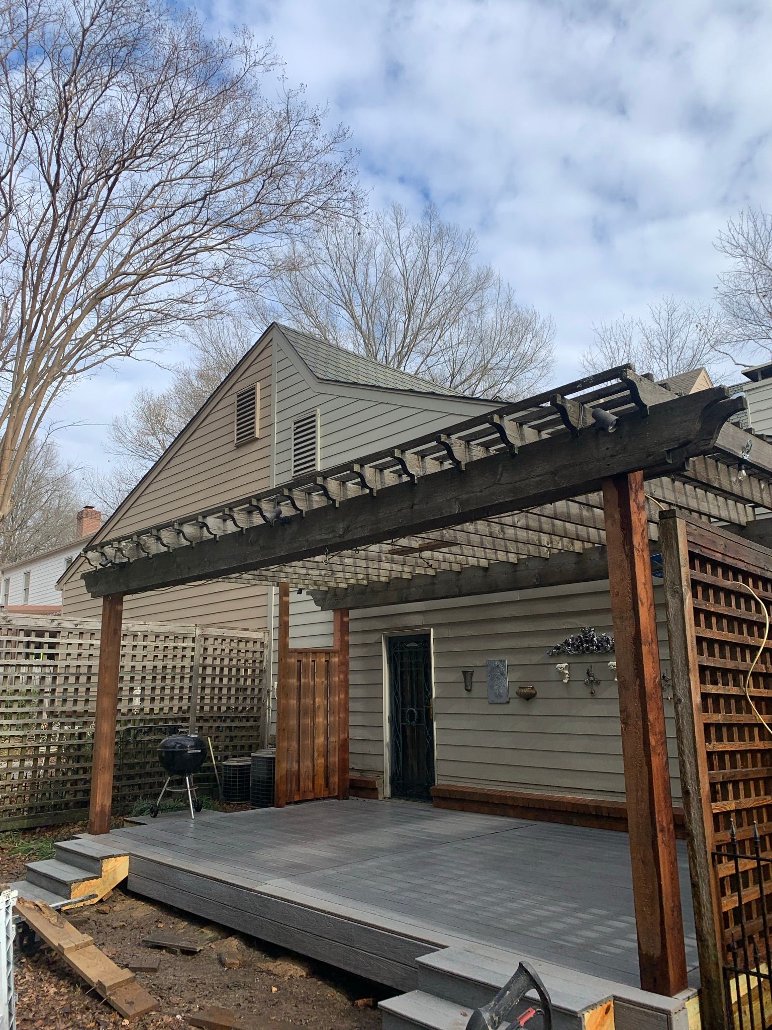 Wooden pergola over a deck with a house in the background.