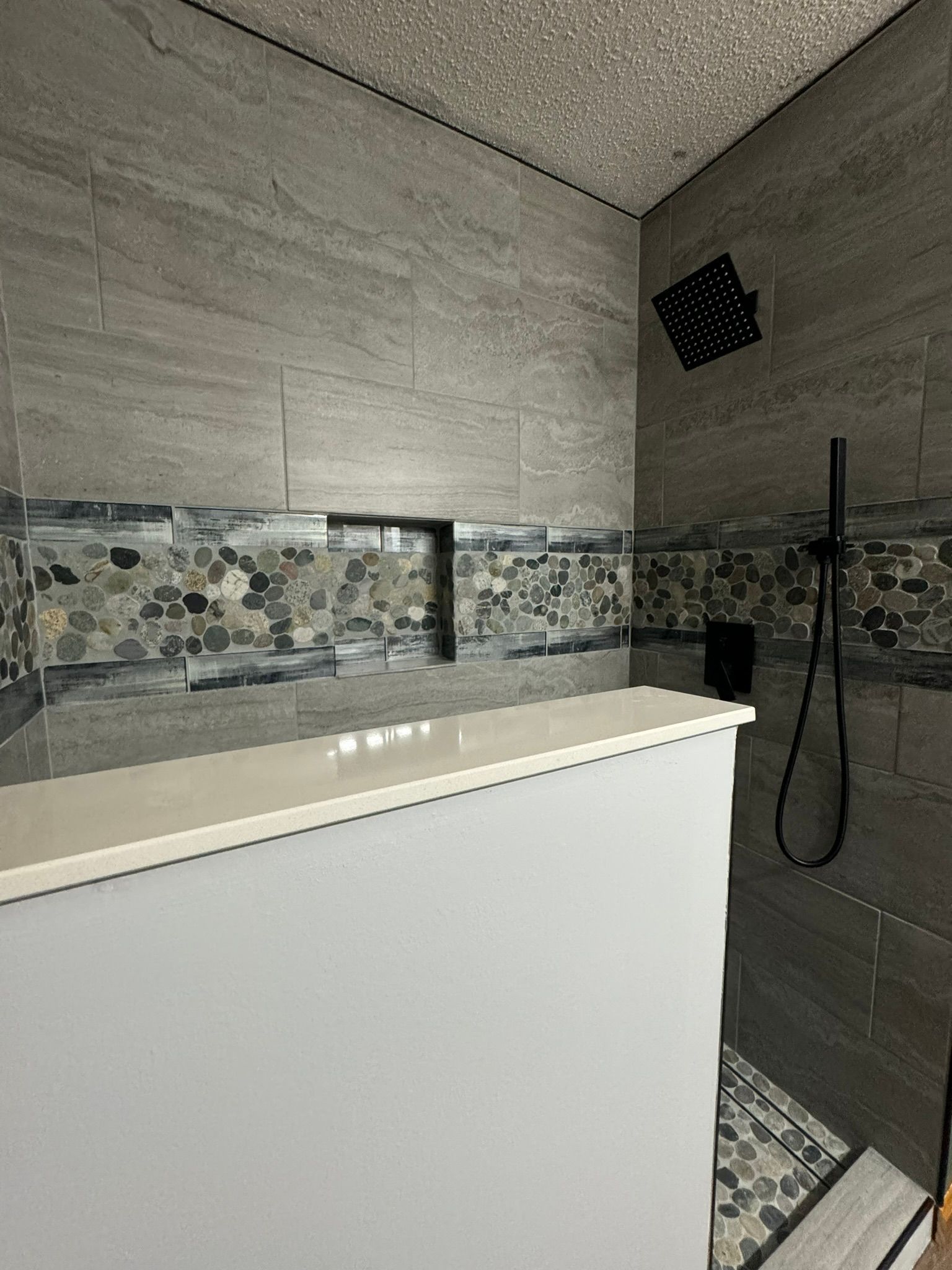 Gray tiled shower with a built-in shelf, black fixtures, and pebble accent border.