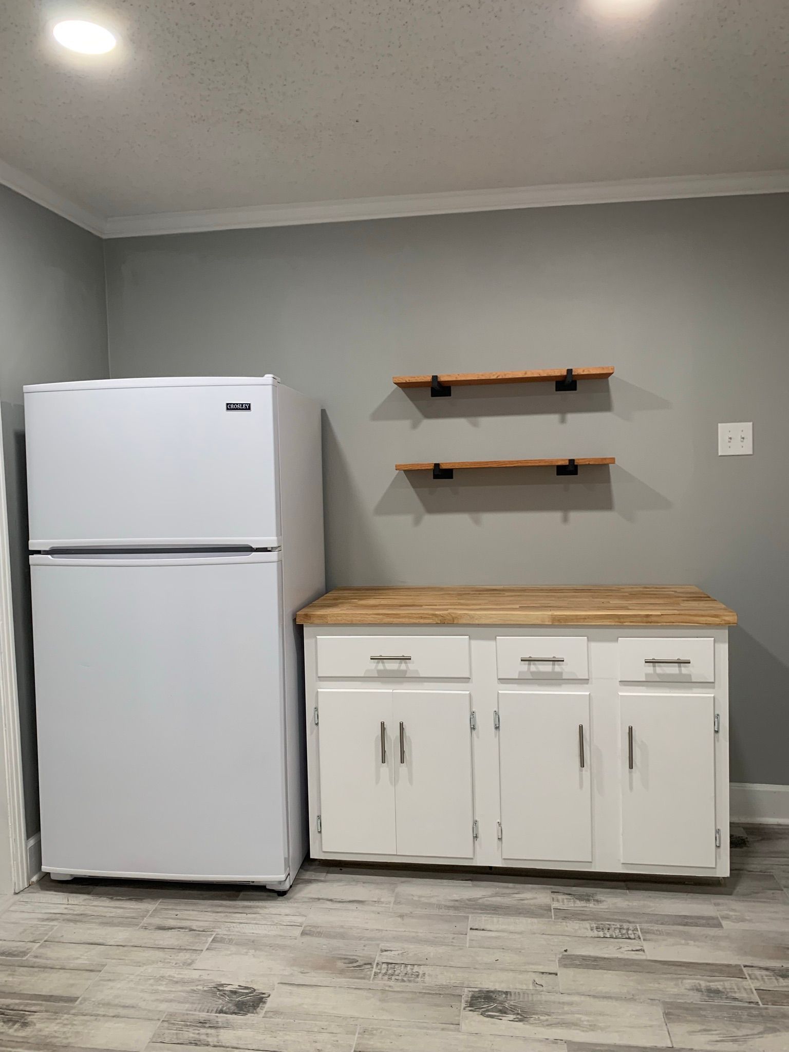 White refrigerator next to white cabinets with a wooden countertop and shelves on gray wall.
