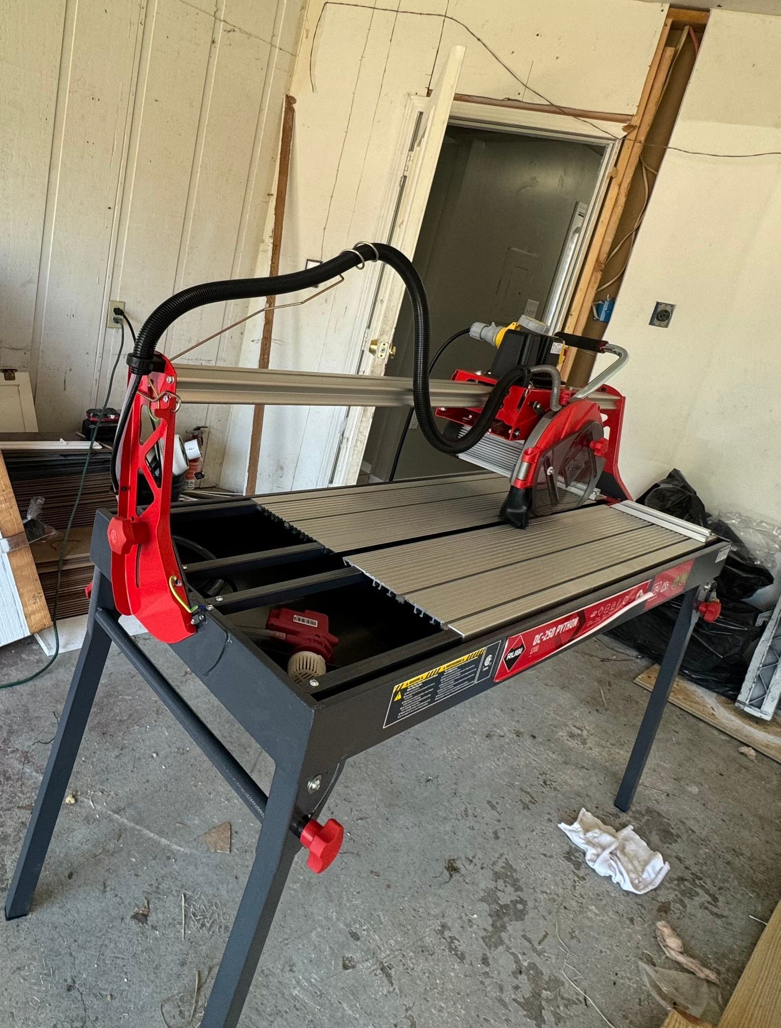 A red and black tile saw on a stand, in a cluttered indoor space.