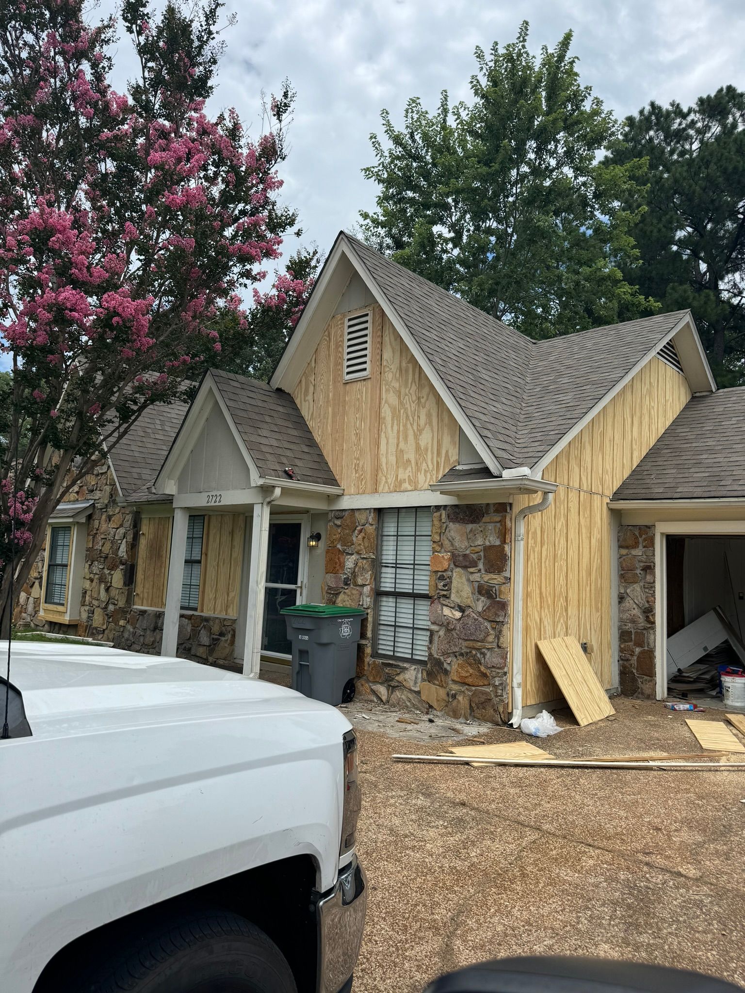 House exterior during renovation with exposed wood siding. Brown stone, green trees, grey sky.