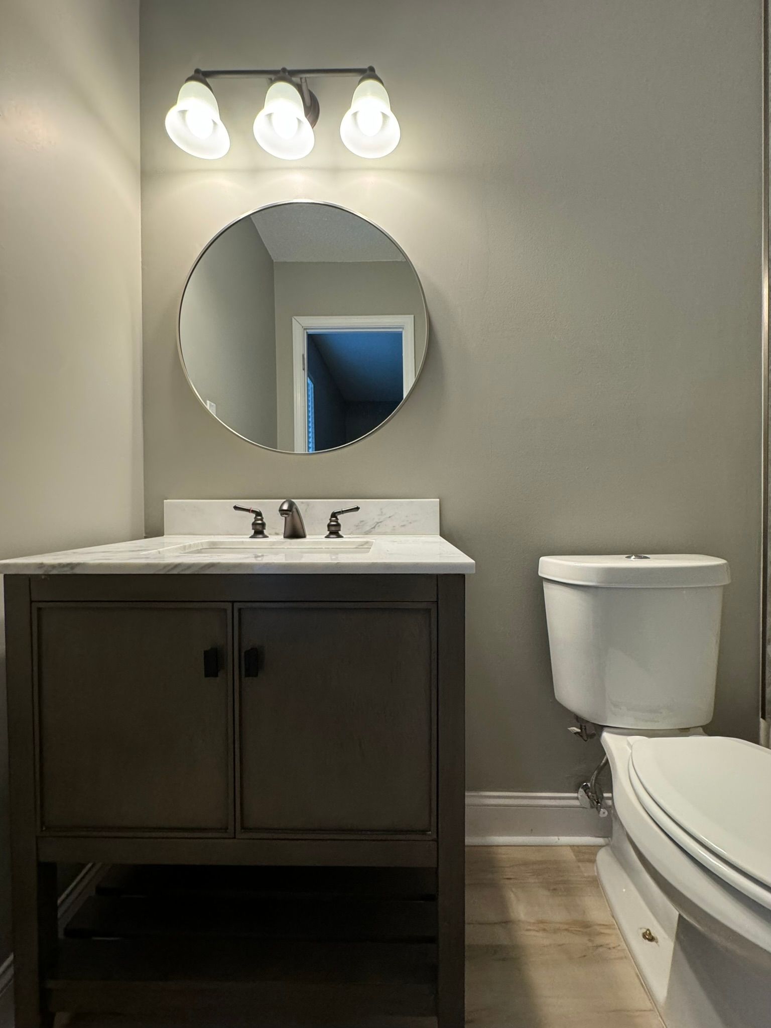 Bathroom with a gray vanity, round mirror, and white toilet. Beige walls and light fixture.