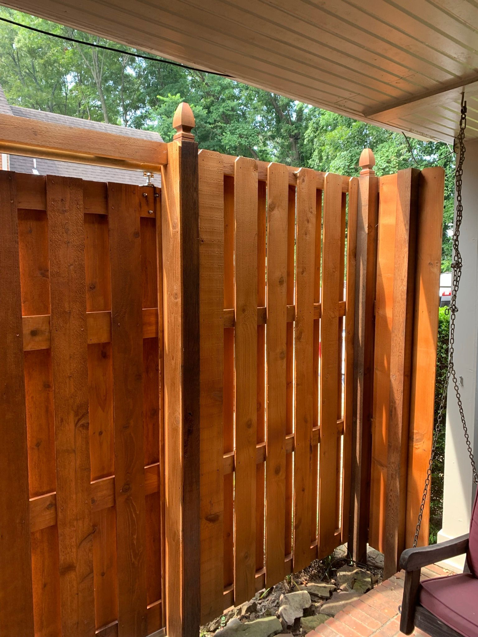Wooden fence with a gate, stained brown, next to a porch. Sunlight.