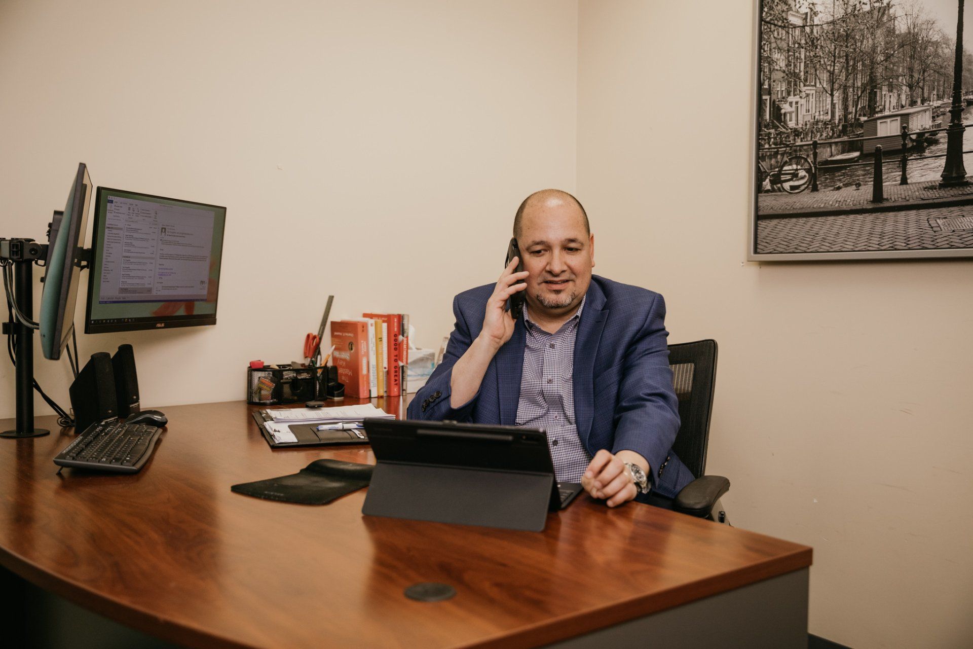 Man in a blue blazer on the phone, seated at a desk with dual monitors and a tablet, in an office.