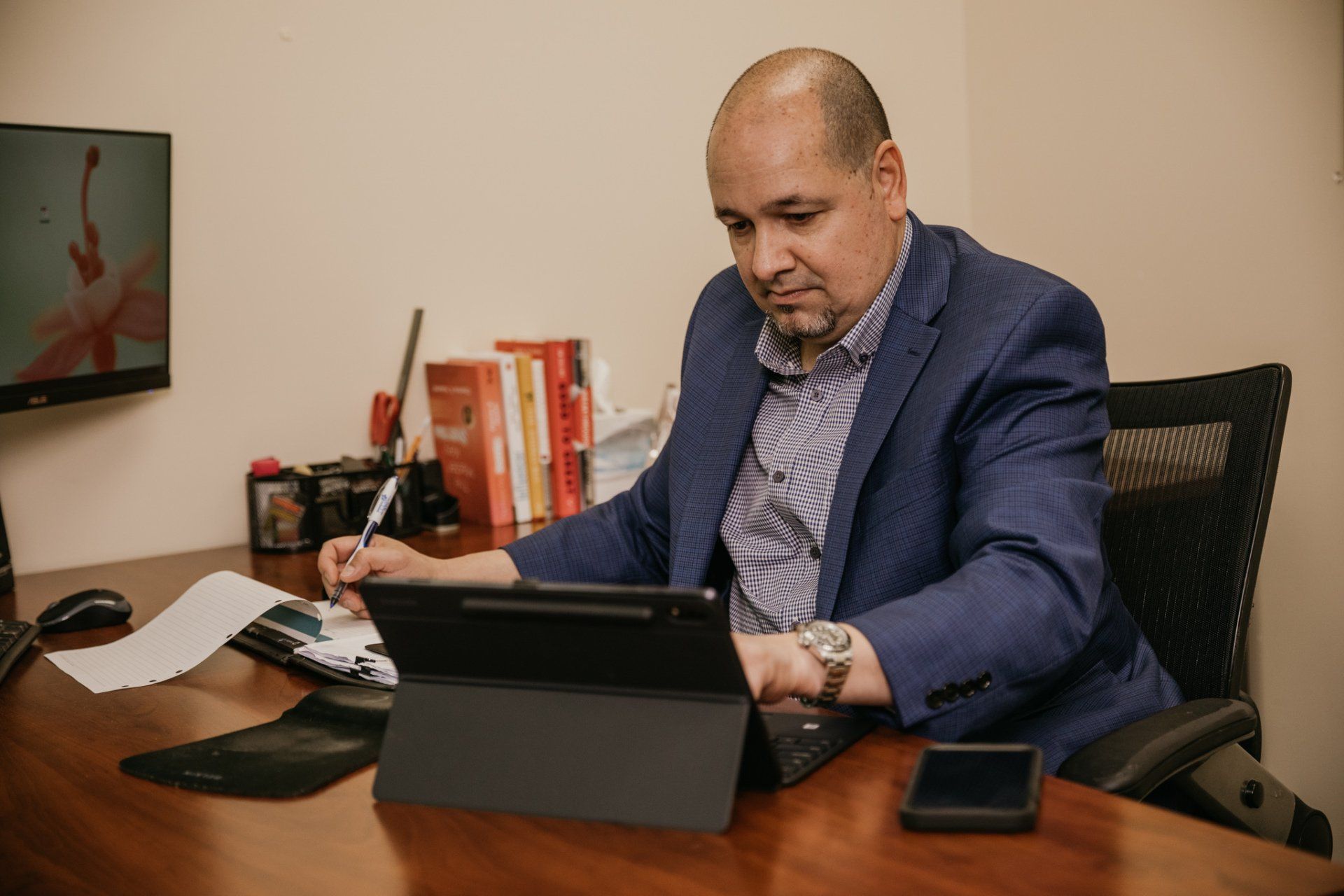 Man in blue blazer at a desk, working on a tablet and writing. Office setting.