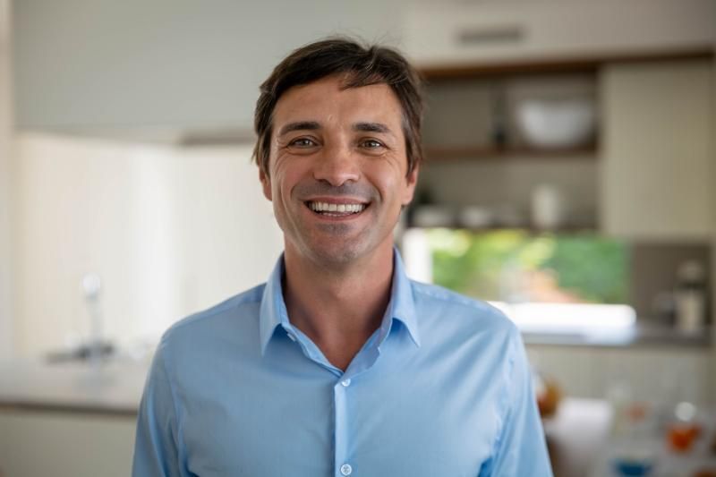 Man smiling indoors, wearing a light blue button-down shirt. Blurred kitchen in background.