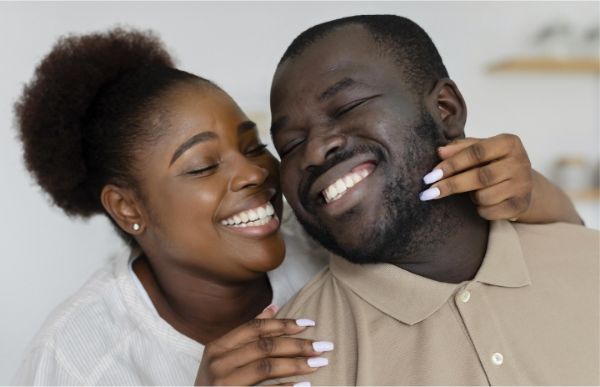 Woman hugs man, both smiling with closed eyes. Light-skinned background.