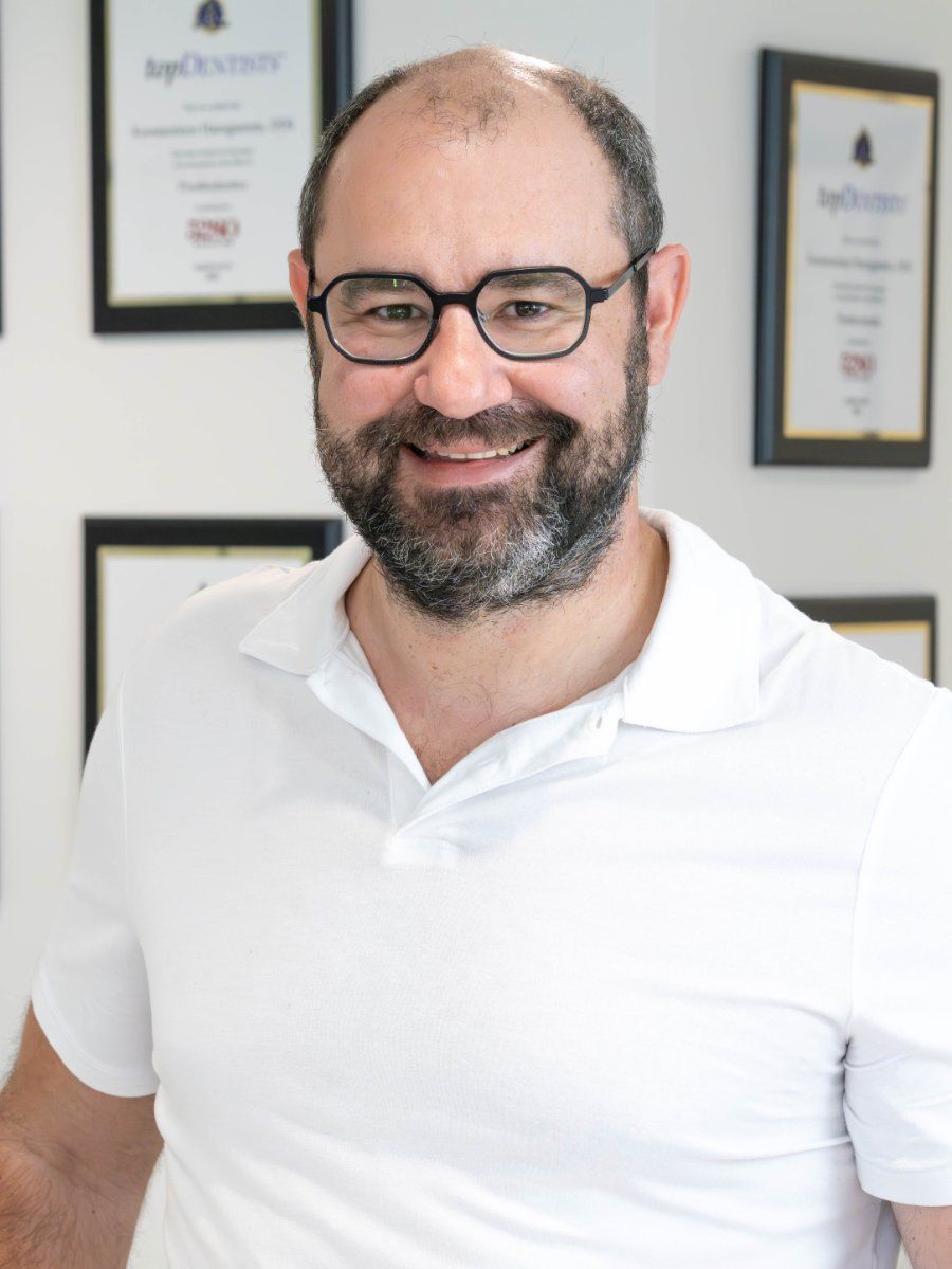 Man with glasses smiles, wearing white polo. Background: framed certificates.