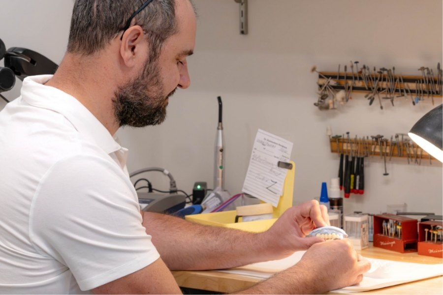 A man with a beard working on a dental mold at a workbench, under a lamp.