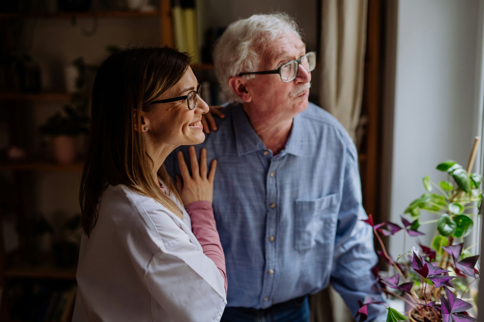 An elderly man and a nurse are looking out of a window.