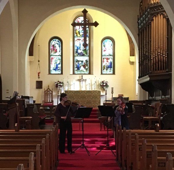 Two musicians playing violins in a church, facing a stained-glass window and cross. Red carpet, pews visible.