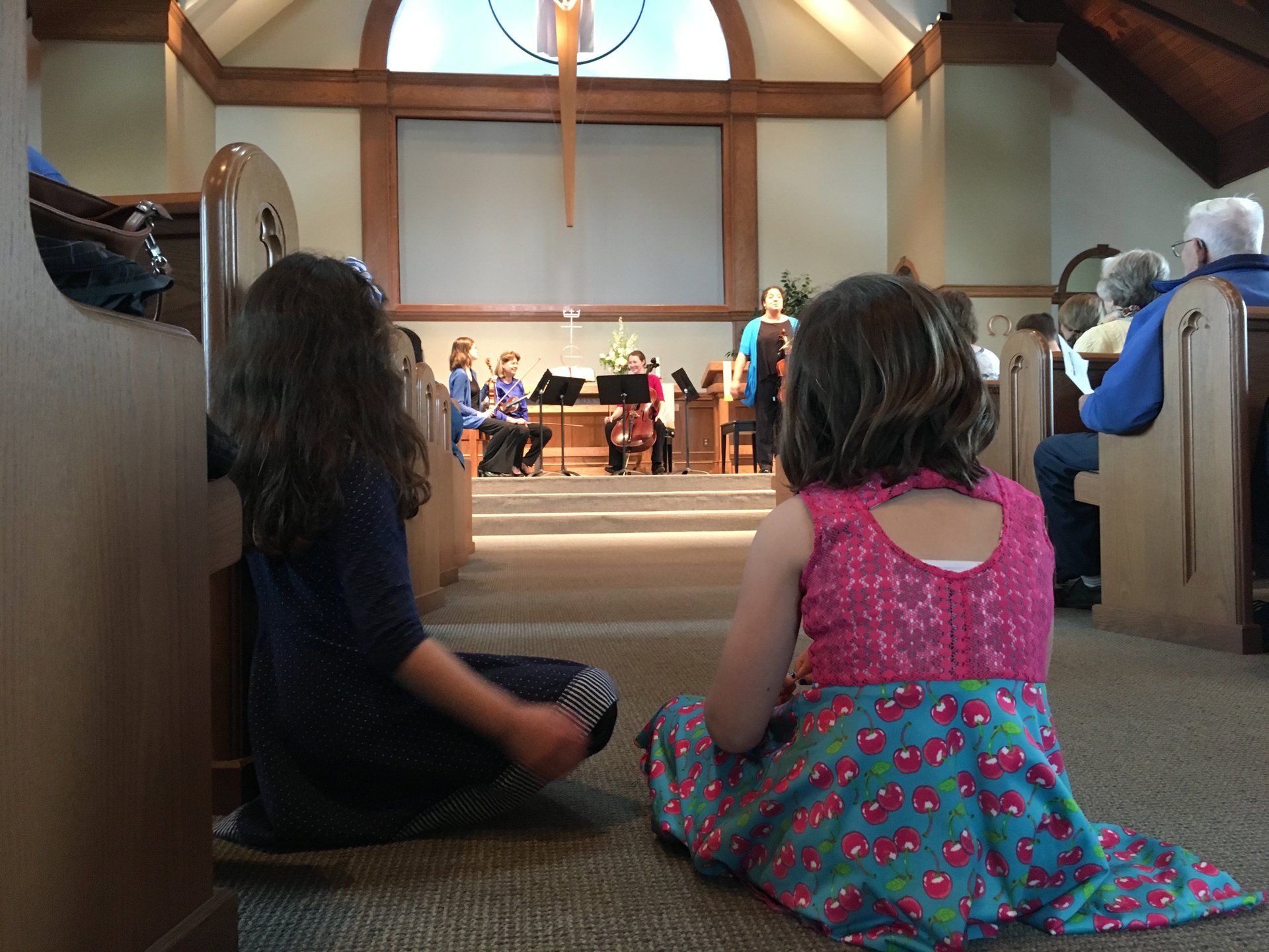 Two children sit on the floor of a church, watching musicians on the stage.