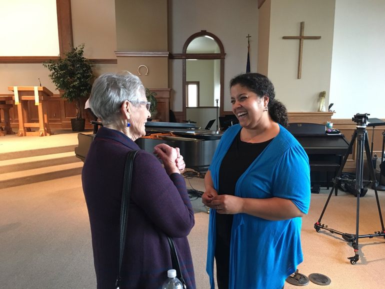 Two women smiling, chatting inside a church. One in purple, the other in blue, both facing each other. Cross visible on the wall.