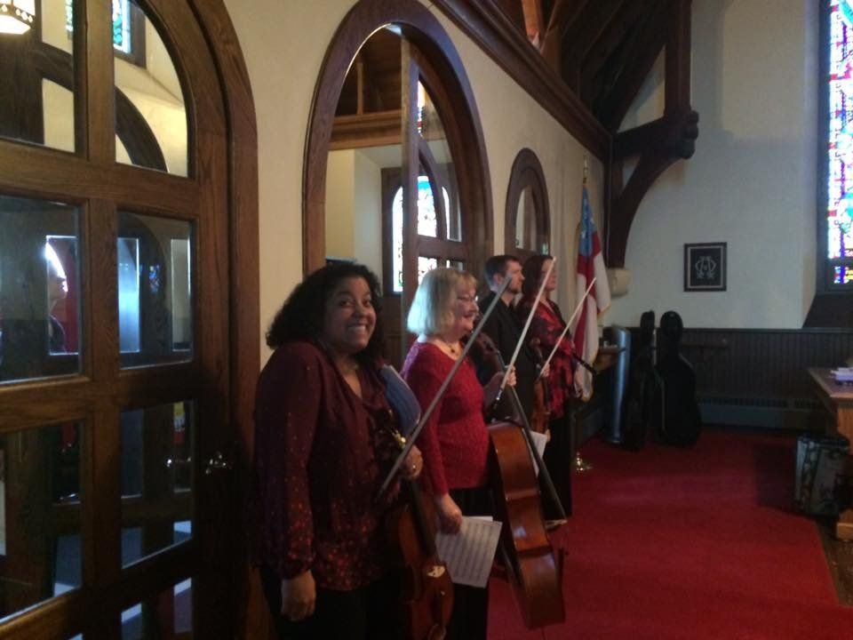 Musicians with stringed instruments in a church, red carpet, stained glass window.