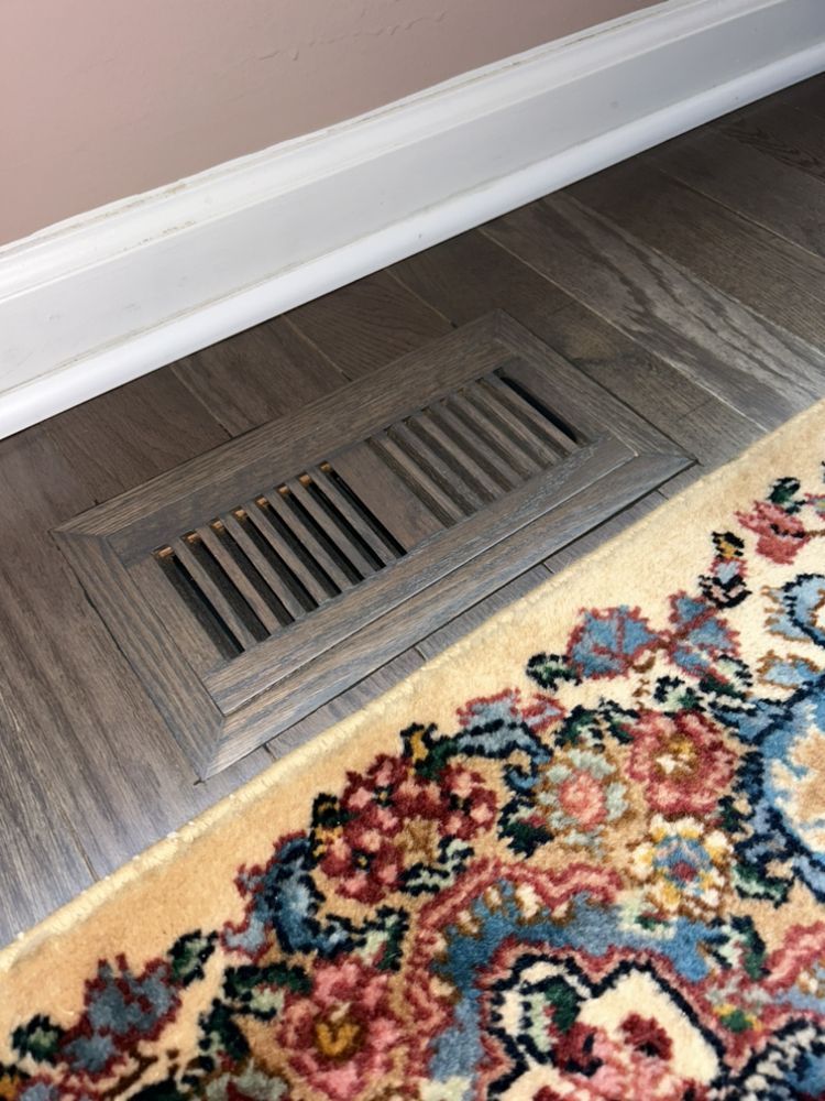 Floor vent, partially covered by patterned rug, near pink wall and white trim.