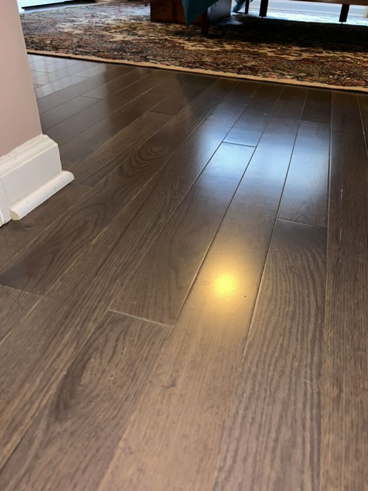 Dark gray hardwood floor with light reflections; near a white baseboard and patterned rug.