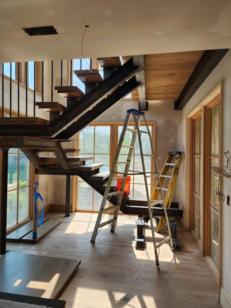 A staircase under construction with metal supports, wooden steps, and two ladders in a room with large windows.