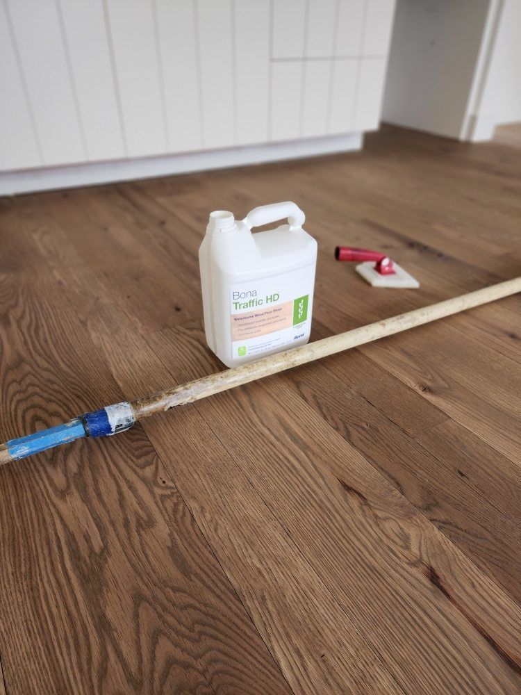 A can of floor finish, applicator, and tools on a newly finished hardwood floor.
