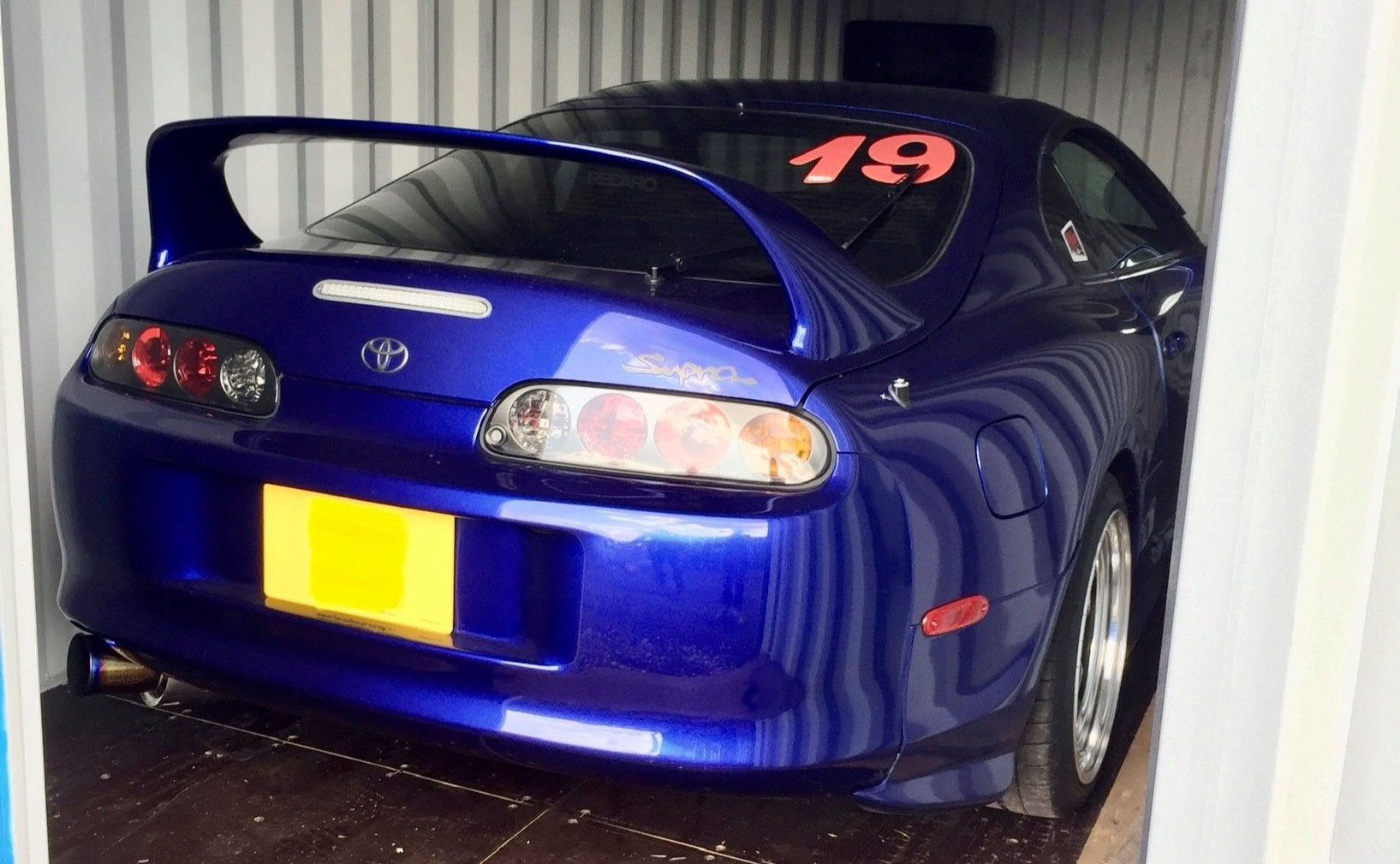 A deep blue Toyota Supra with a racing number 19 on the rear window, parked inside a metal storage container.