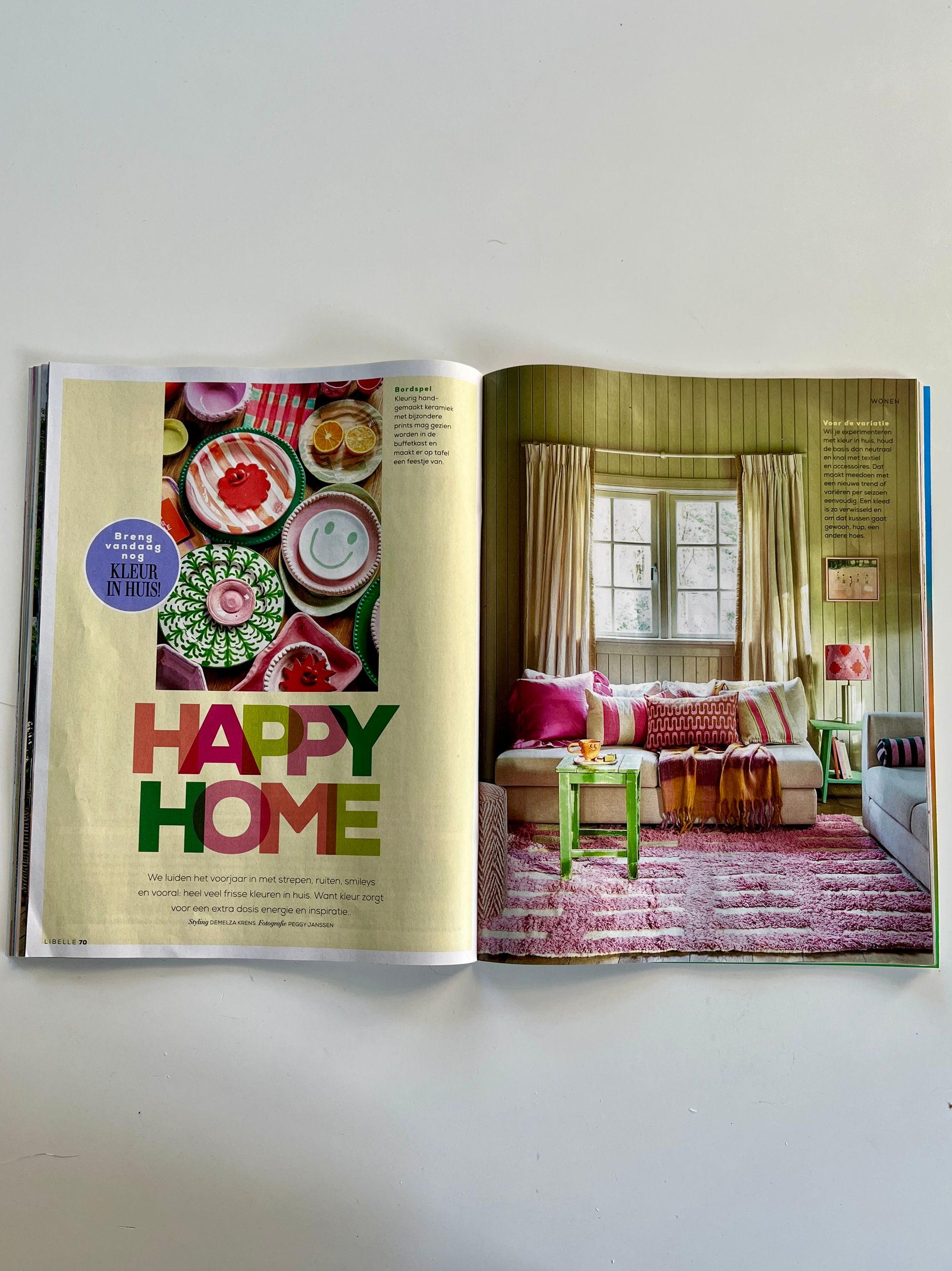 Een paginagrote afbeelding uit een tijdschrift van een kamer met een roze vloerkleed met patroon, een crèmekleurige bank, een groene bijzettafel en een collage van kleurrijke borden.