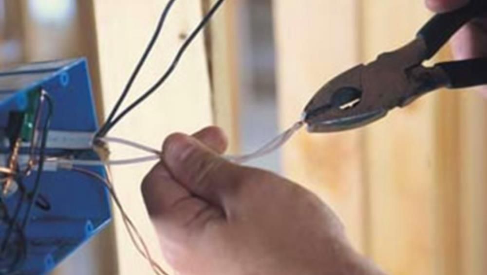 Person cutting a white electrical wire with pliers near a blue electrical box.