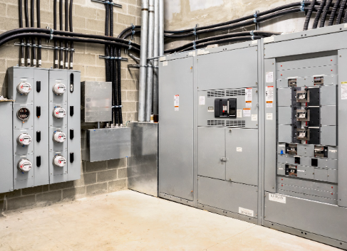 Electrical room with gray metal panels, meters, conduits, and wiring on brick walls.