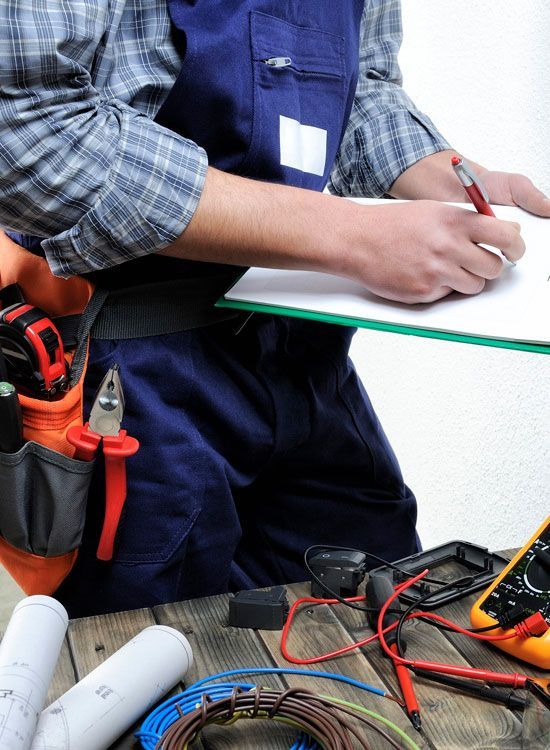 Electrician writing on clipboard, tools in belt, wiring and multimeter on table.