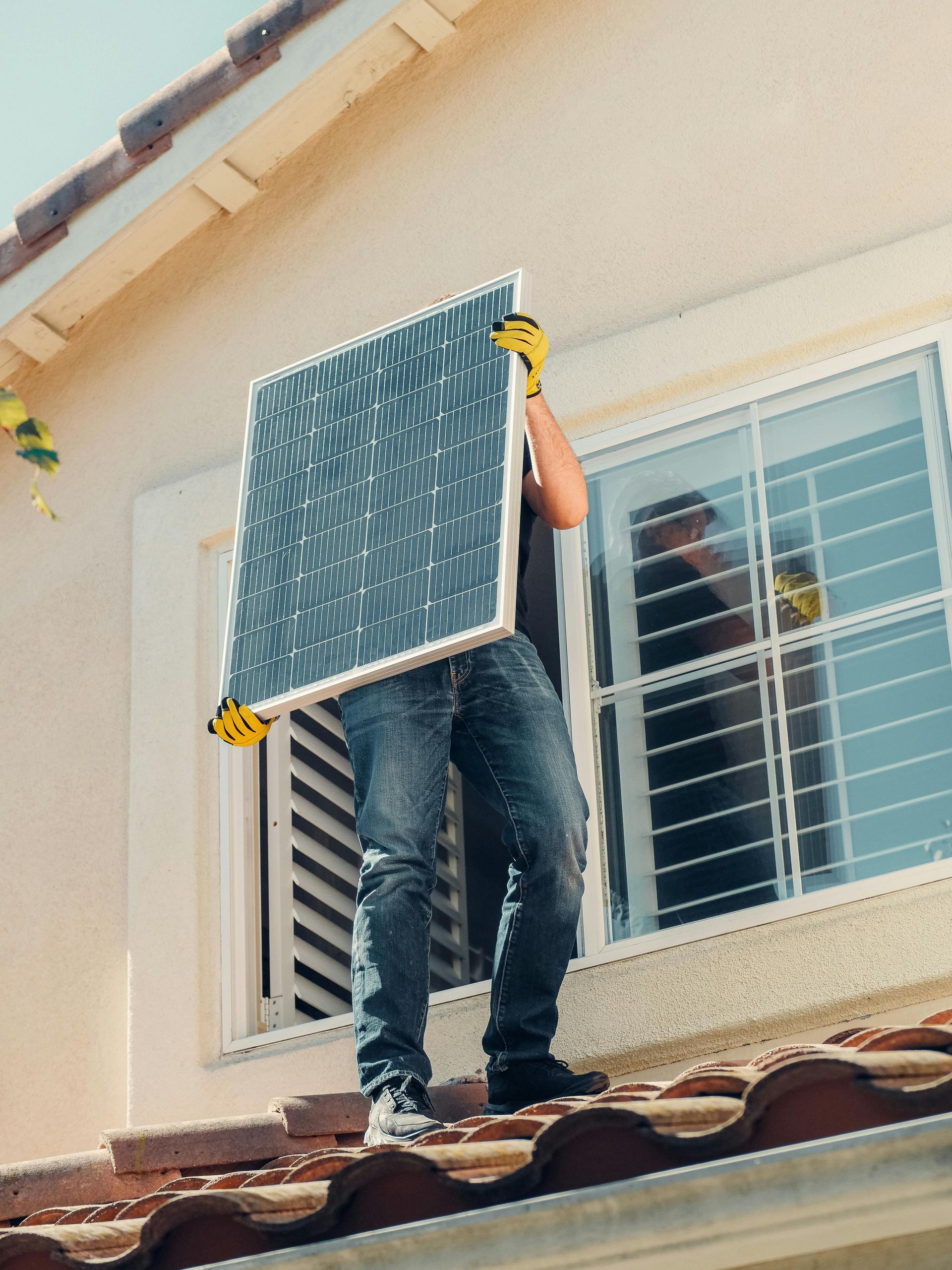 Person holding a solar panel on a rooftop near a window. Yellow gloves and blue jeans are visible.