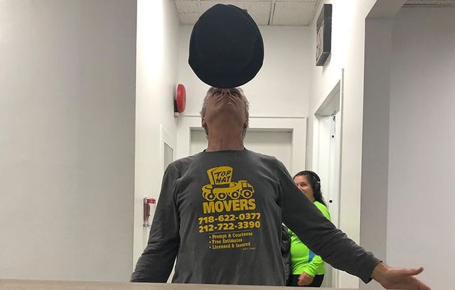 a man wearing a movers shirt is balancing a hat on his head .