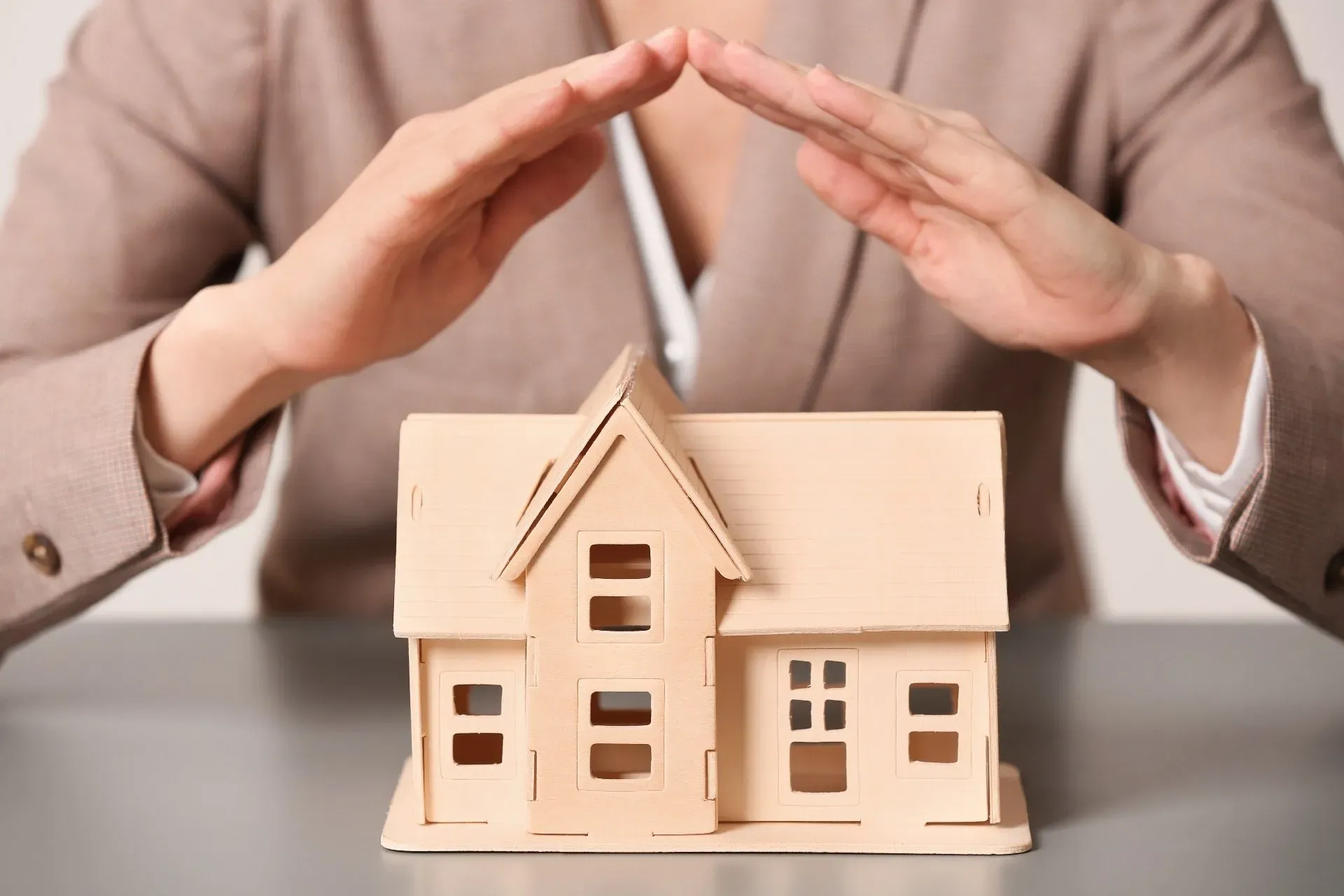 Hands sheltering a miniature house on a desk, symbolizing protection or insurance.