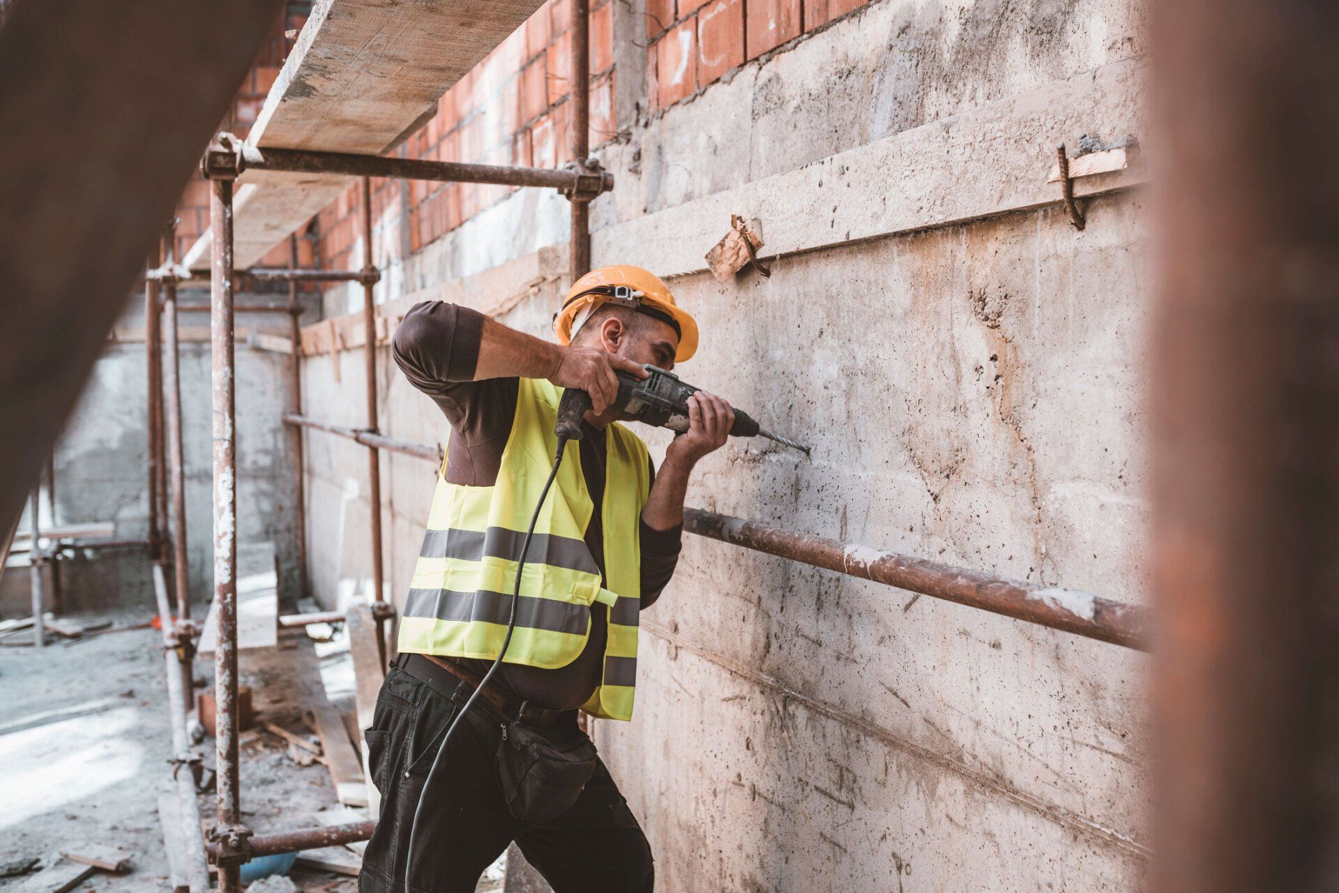 Construction worker drills into a concrete wall, wearing a hard hat and safety vest.