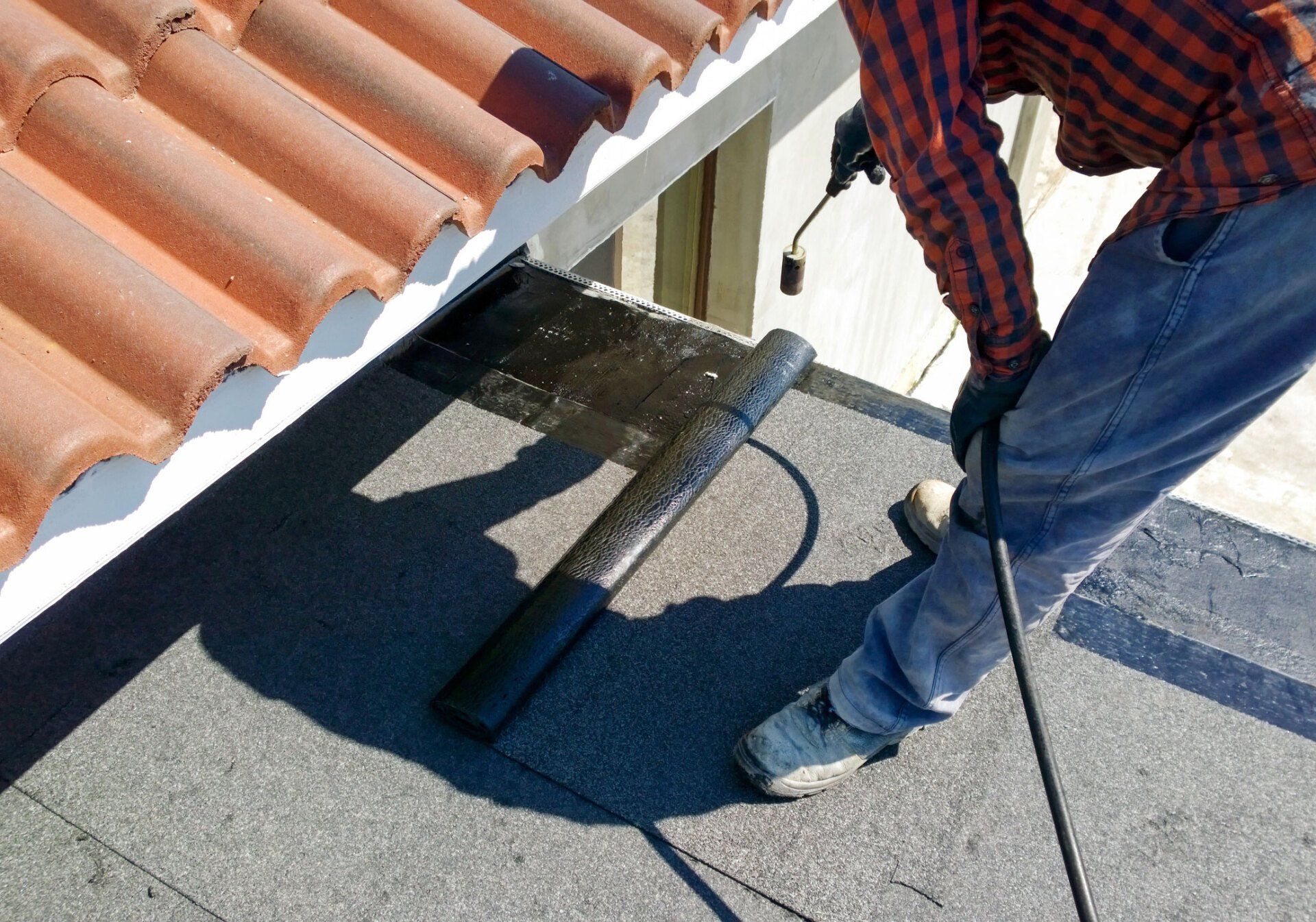 Person using a torch to install roofing material near a tiled roof.