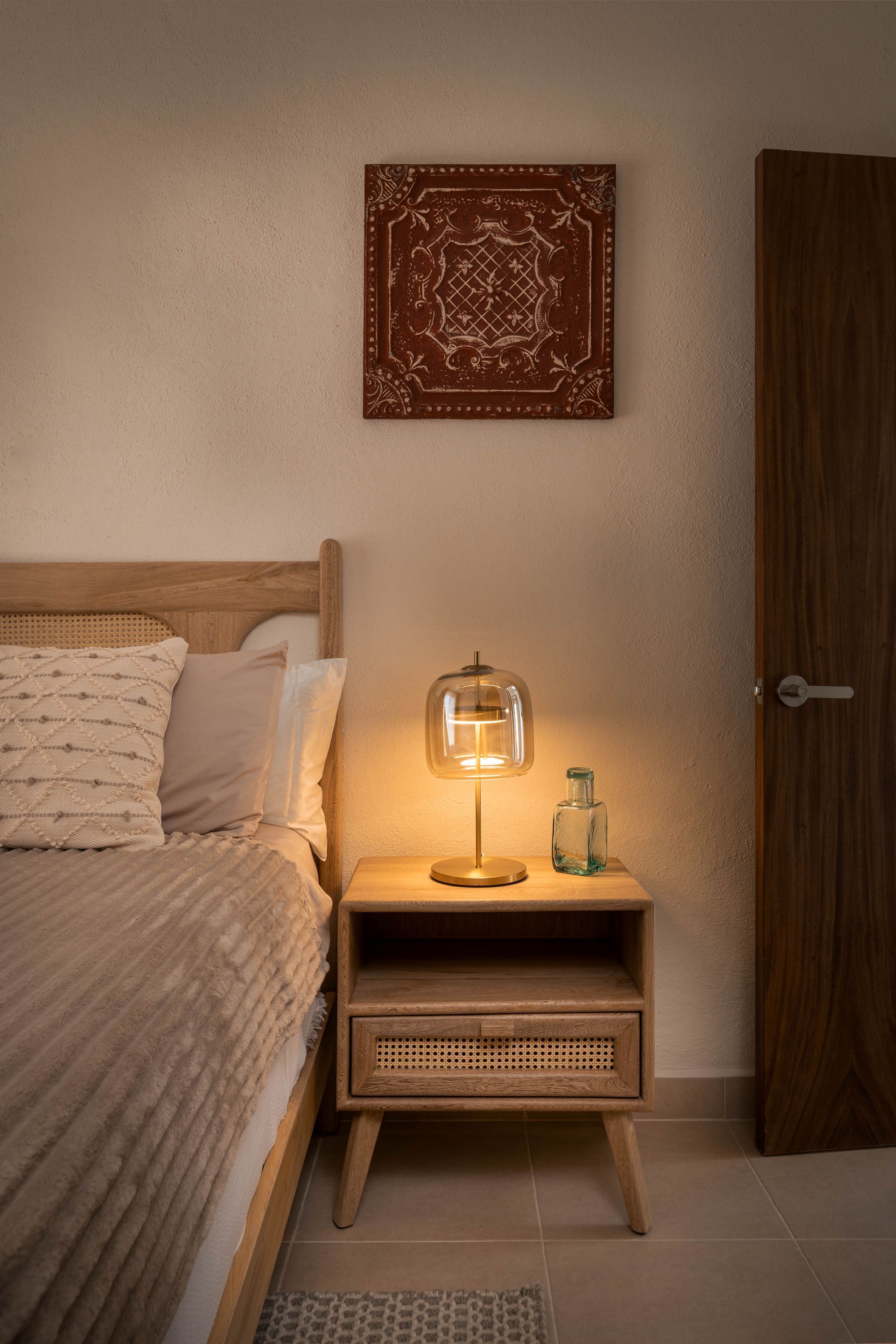 A bedroom with a bed , nightstand , lamp and painting on the wall.