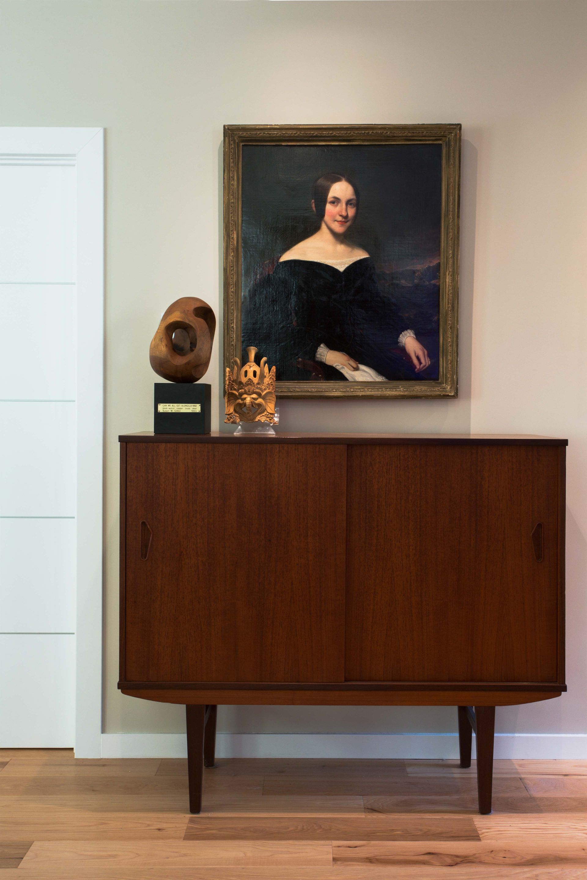 A painting of a woman is hanging on a wall above a wooden dresser.