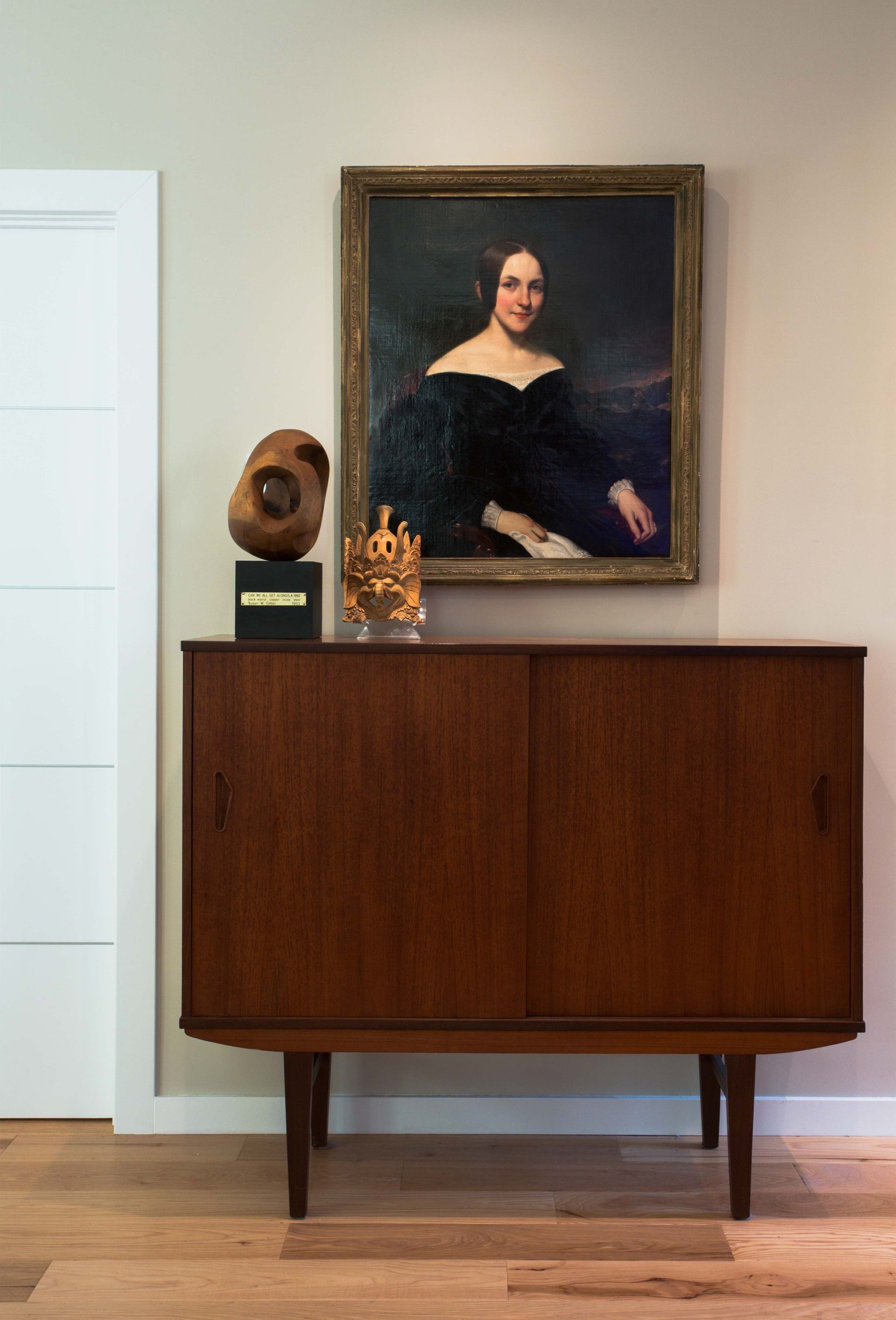 A painting of a woman is hanging on a wall above a wooden dresser.