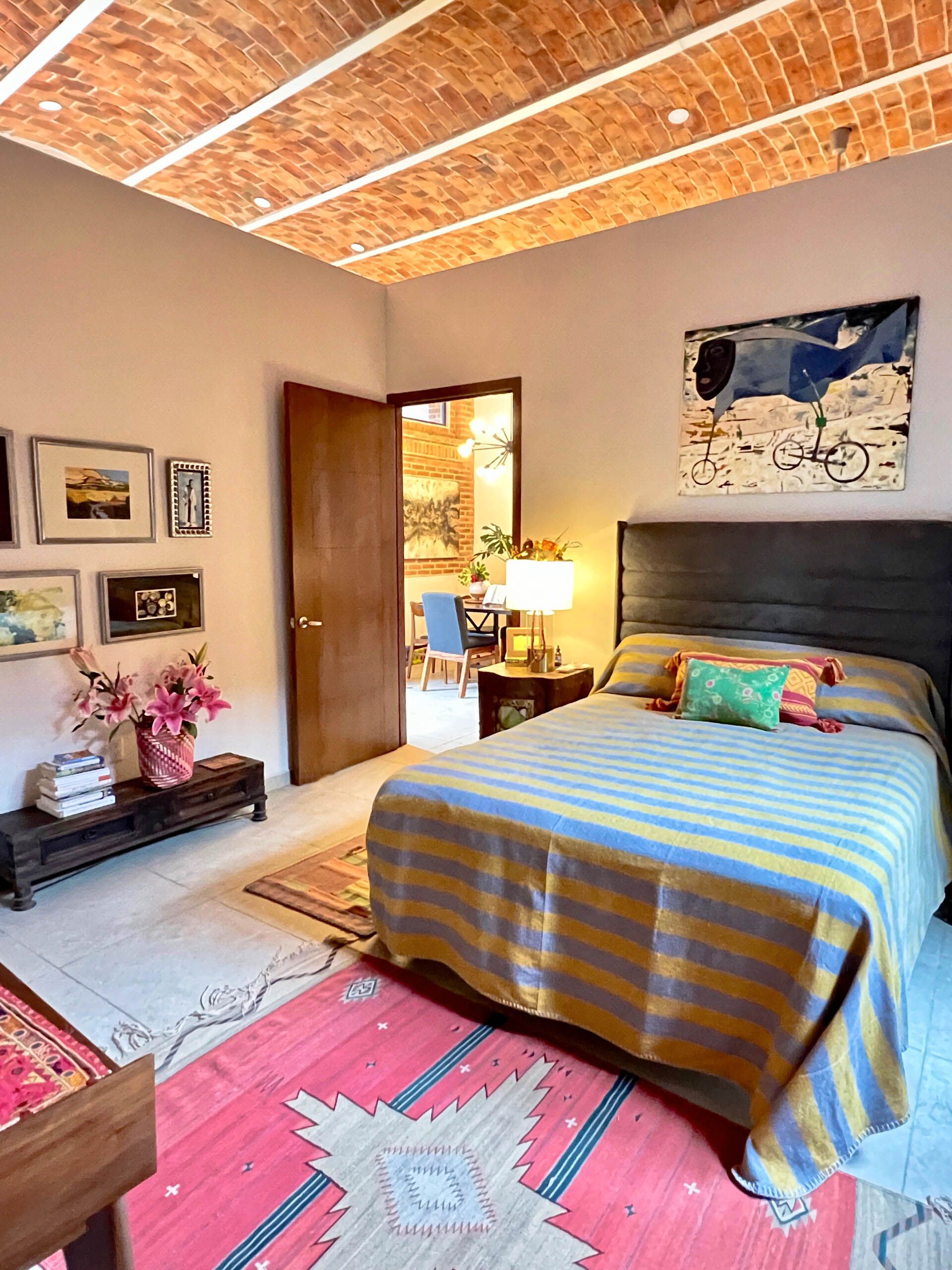A bedroom with a bed , rug , paintings and a brick ceiling.