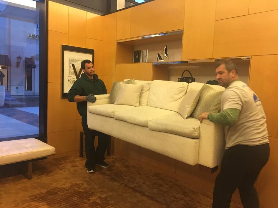 Two men are carrying a white couch in a living room.