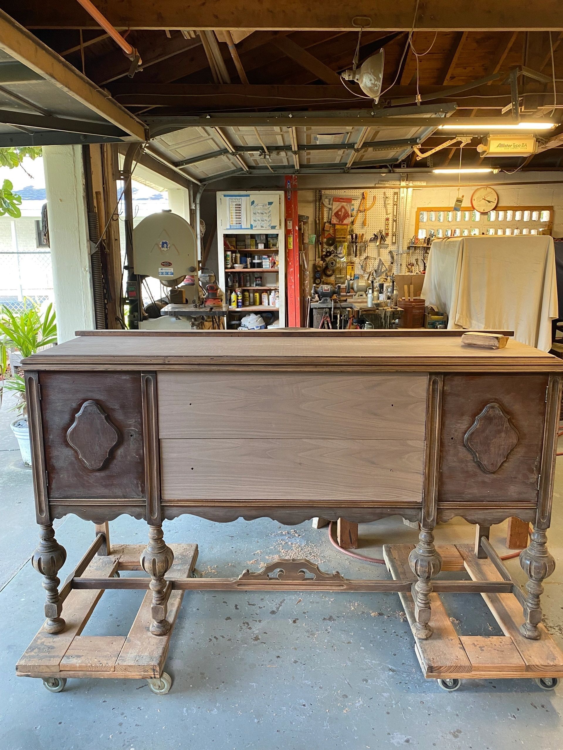 Antique wooden buffet on dollies, partially stripped. Garage setting with tools and equipment visible in background.