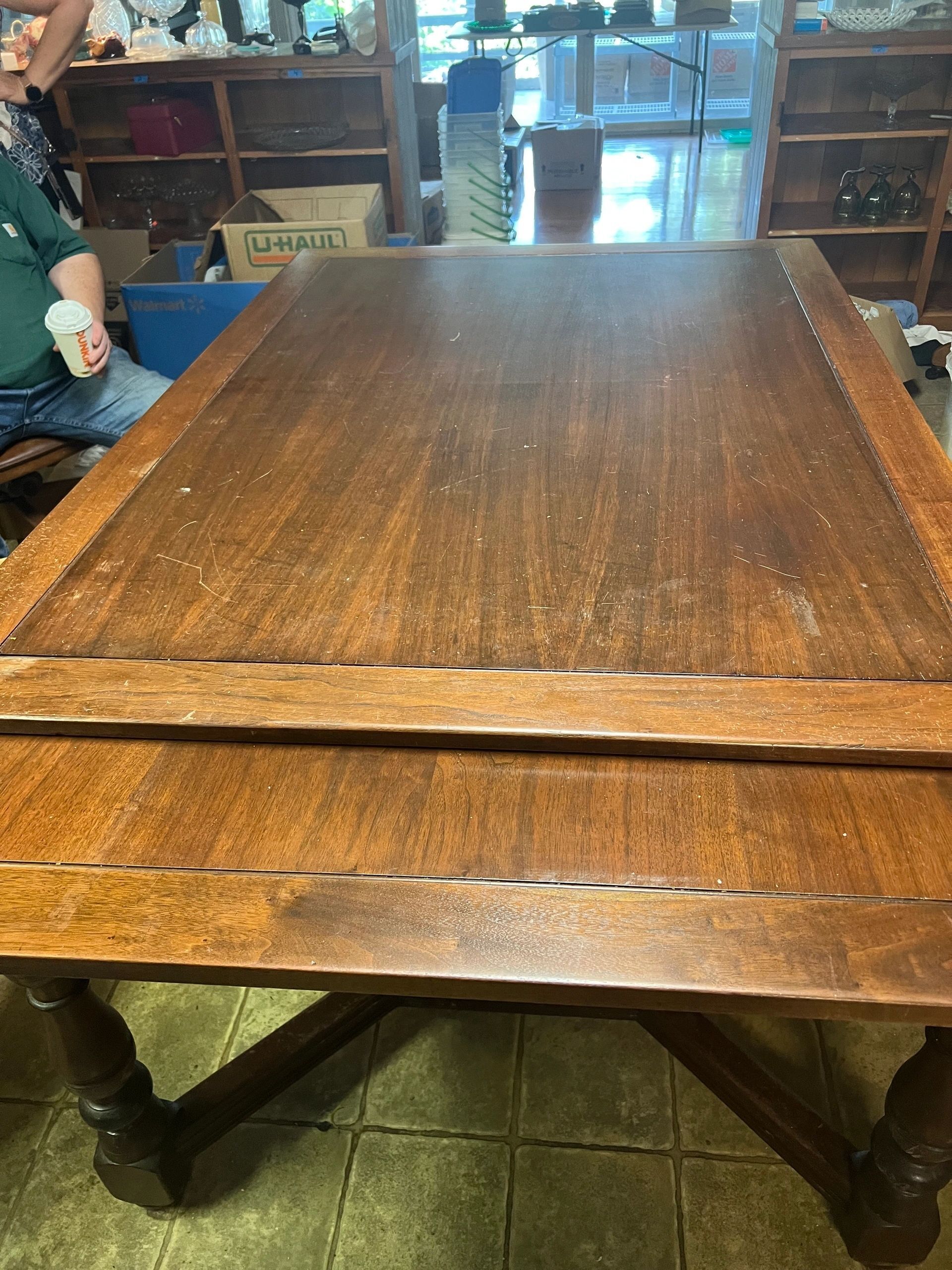Wooden dining table with turned legs, in a room with shelves and a person.