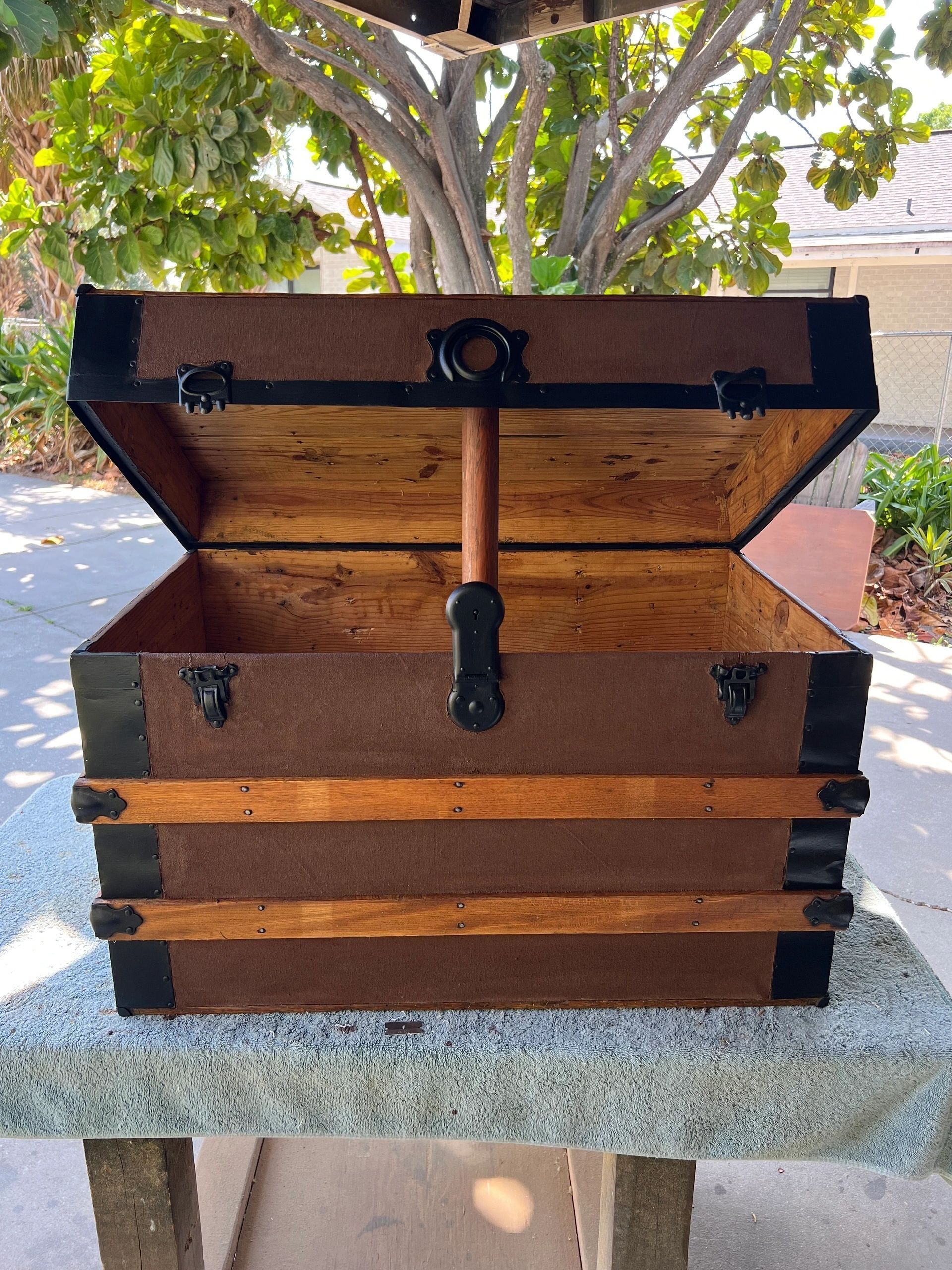Antique brown trunk with black metal trim, lid open, wooden handle, set outdoors on a stone surface.