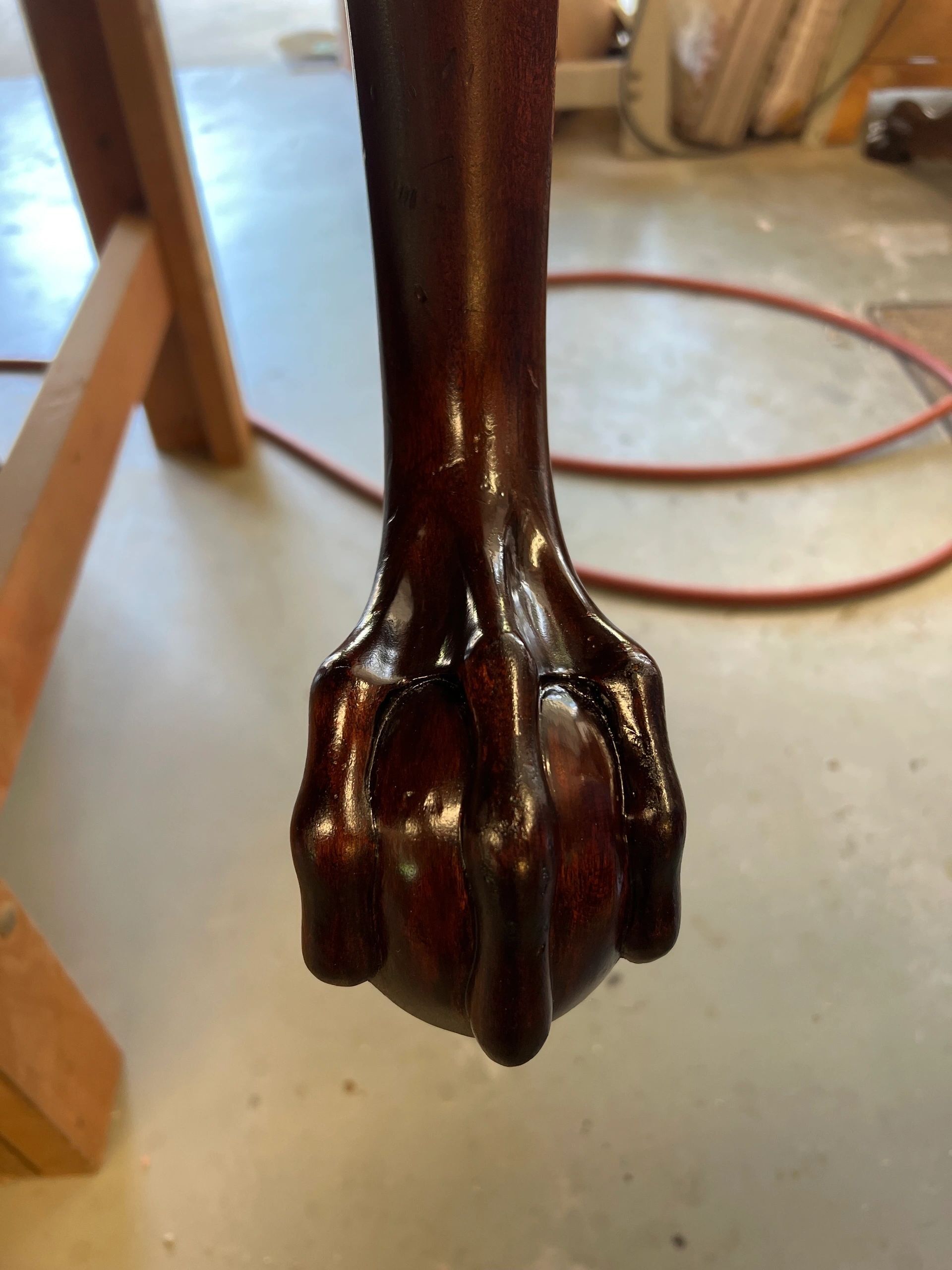 Close-up of a mahogany-colored furniture leg with a carved claw foot. Shiny finish, workshop setting.