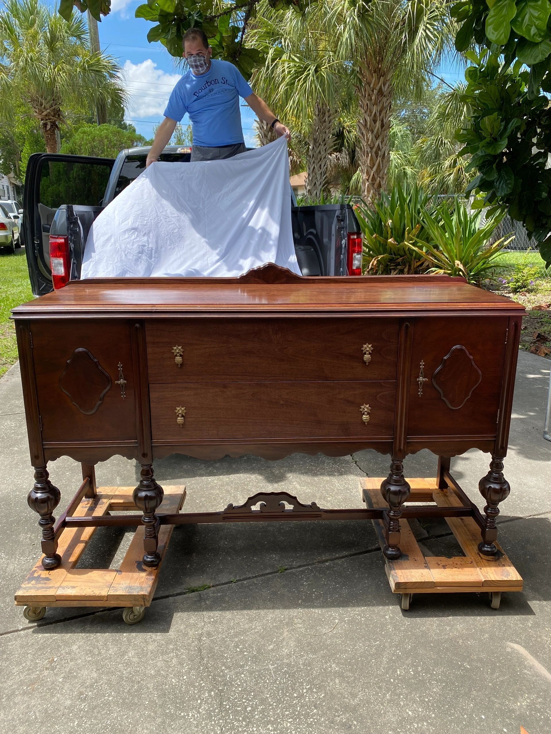 A dark wooden buffet on dollies, being covered with a sheet by a person standing behind a pickup truck.