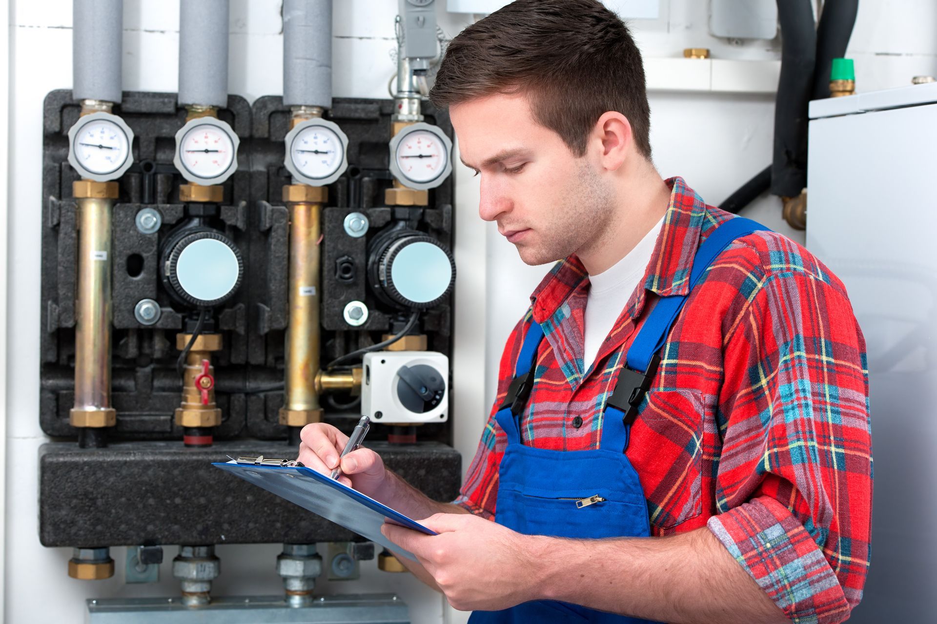 Technician checks heating system gauges and controls, wearing plaid shirt and blue overalls.