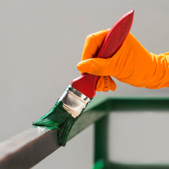 Mano en guante naranja pintando una barra de metal verde con un pincel.