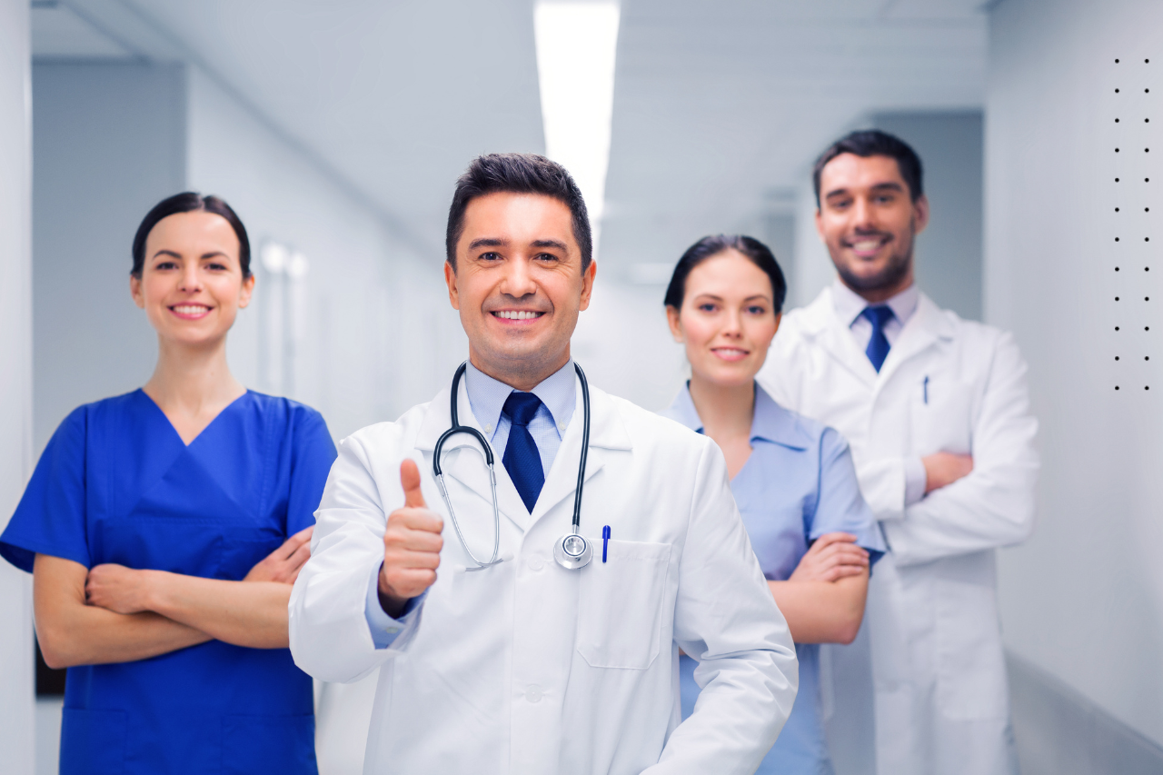 Four medical professionals in a hospital hallway; smiling, one giving thumbs up.